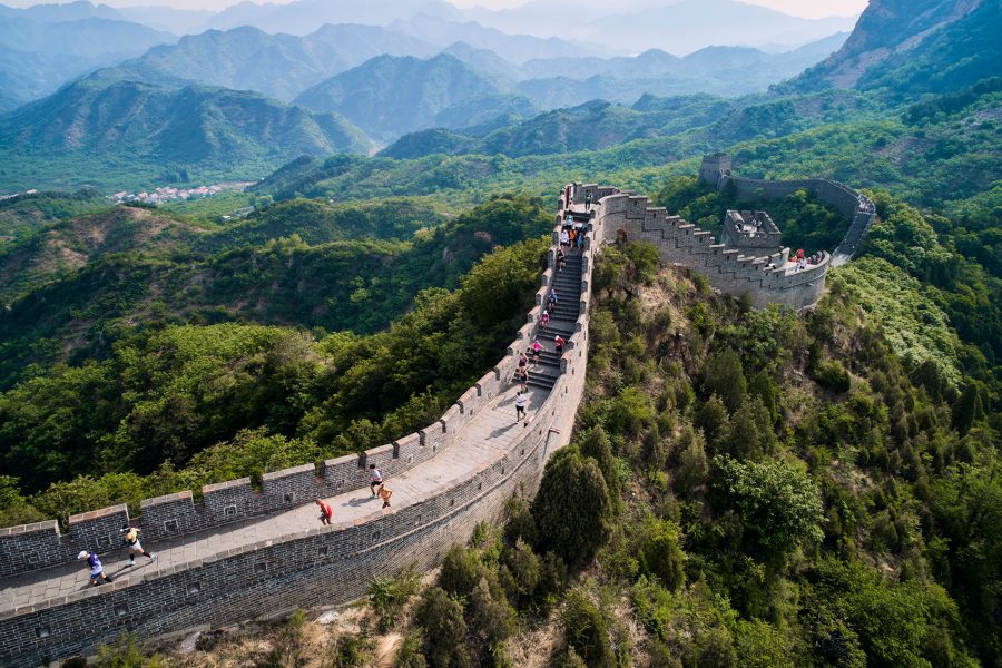 A drone shot of runners running along the Great Wall of China in Beijing surrounded lush green forests and mountains in the distance.