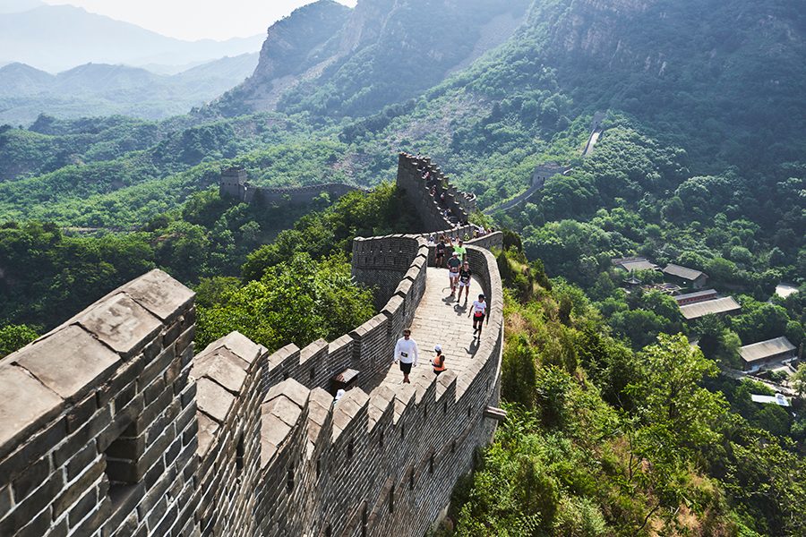 A drone shot of marathon runners climbing up the Great Wall of China on a sunny day, surrounded by lush green forests and mountains.
