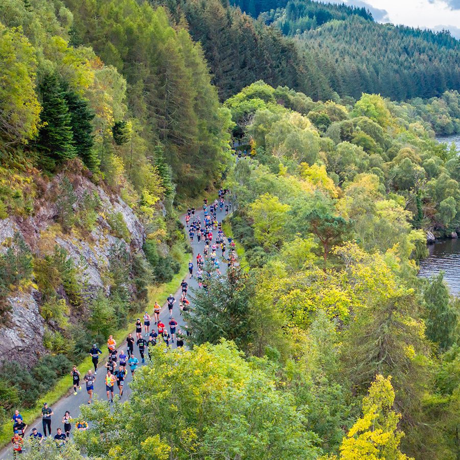 A drone shot of a crowd of runners along the Scottish Highlands on the Loch Ness Marathon waterside route with a view of the lake and mountains