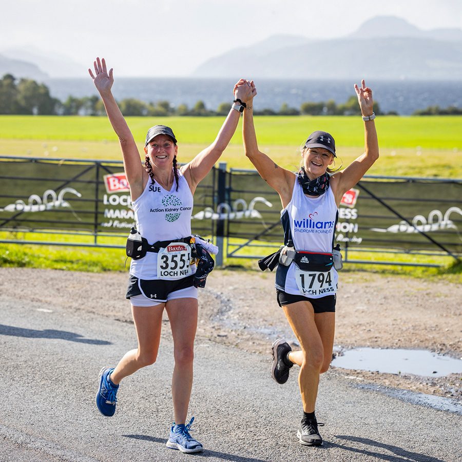 A pair of women runners smiling, cheering and jogging on the Loch Ness Marathon trail, next to green field in the Scottish Highlands