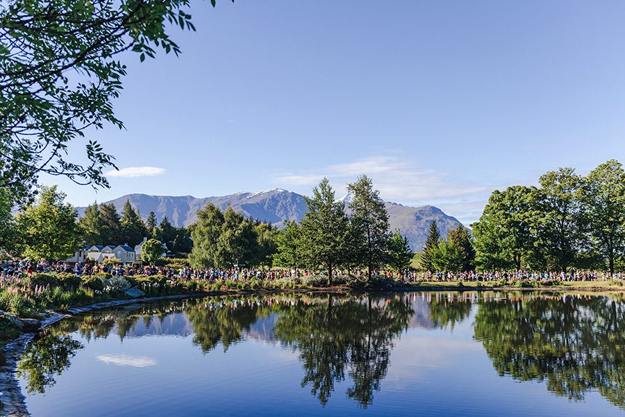 A landscape view of a crowd of marathon runners on New Zealand’s Queenstown Marathon trail by the lake with views of mountains in the background on a clear day