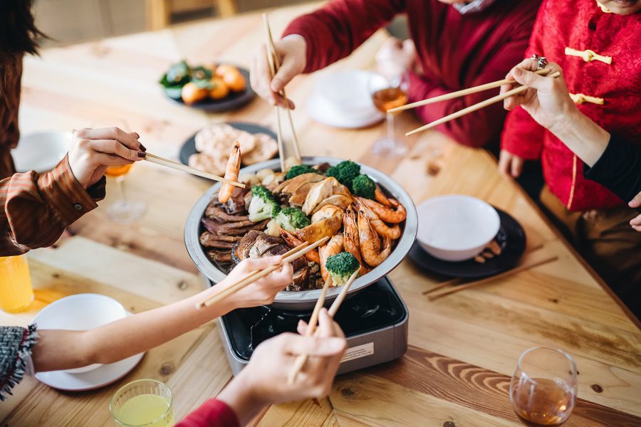 Five pairs of hands holding chopsticks over poon choi on a wooden table.