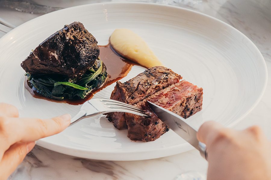  Close-up of hands cutting a piece of steak with a fork and a knife. 