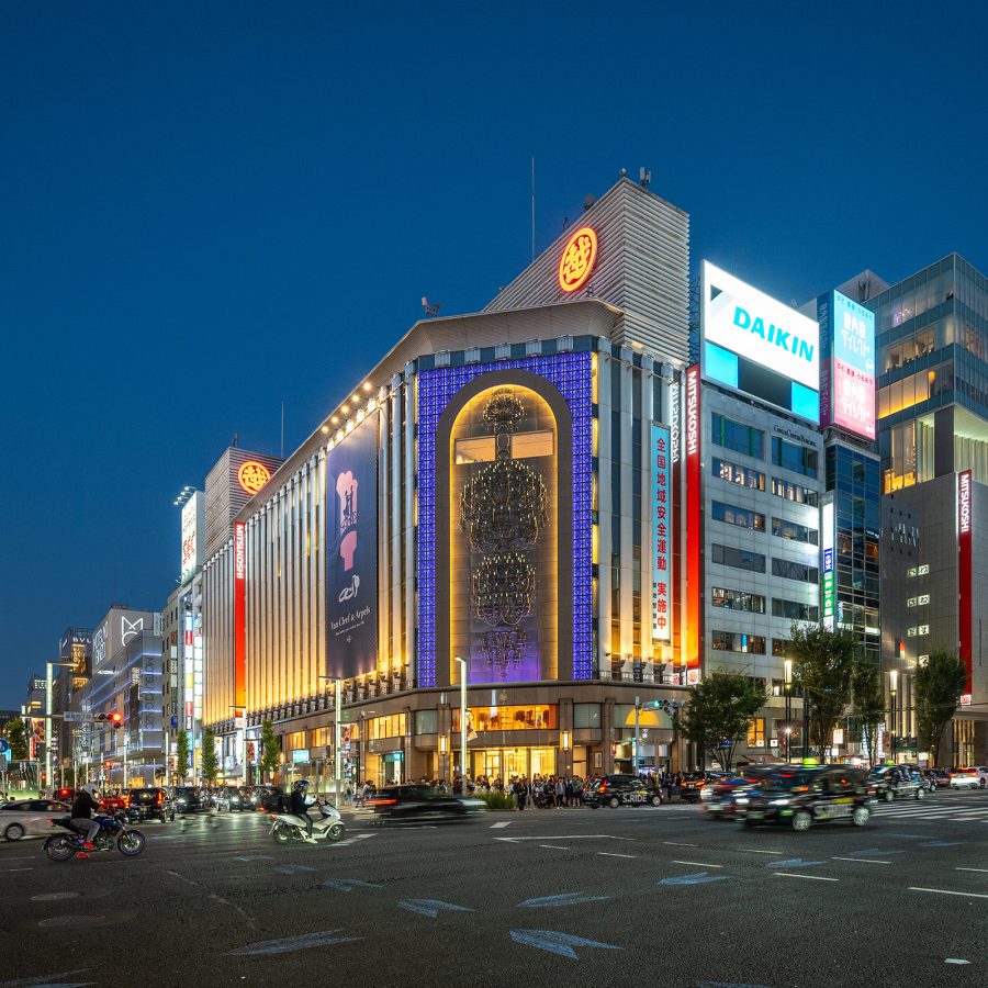 A street view of city lights and people and cars crossing Mitsukoshi Ginza at night.