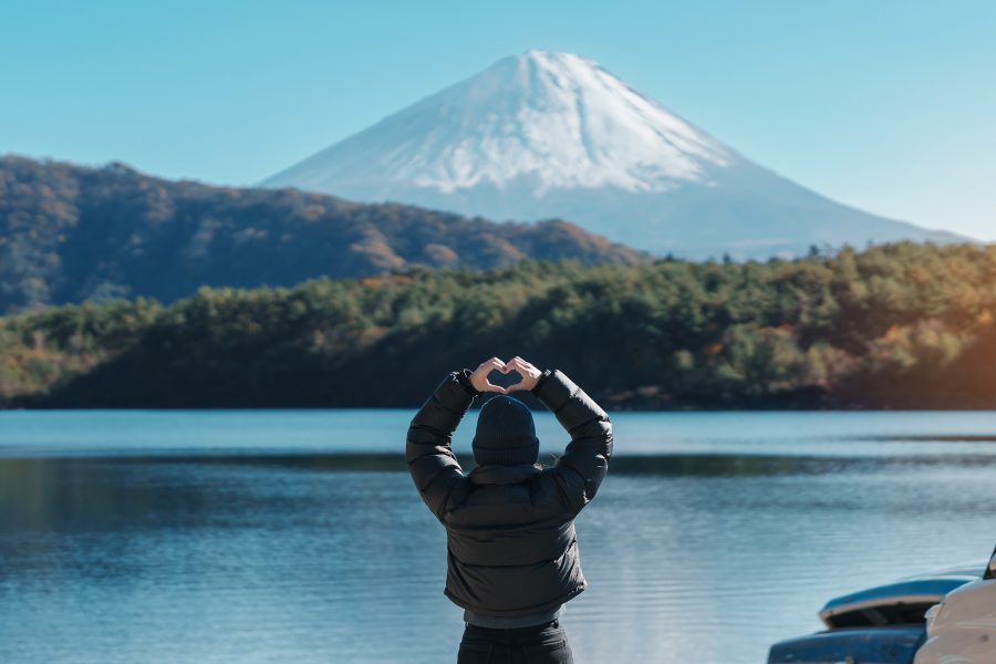 A person seen from behind, standing in front of Saiko Lake holding up a heart shape with their hands, looking towards Mount Fuji in the winter. 