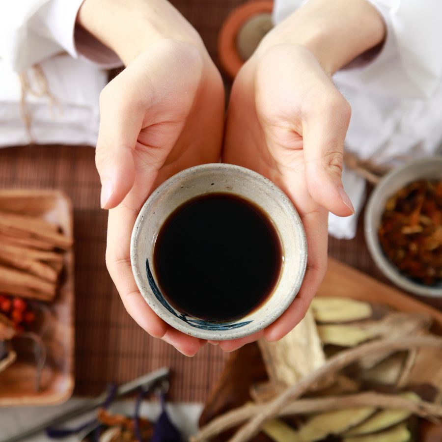 A pair of hands holding a bowl of dark liquid over a table filled with traditional Chinese medicine ingredients. 