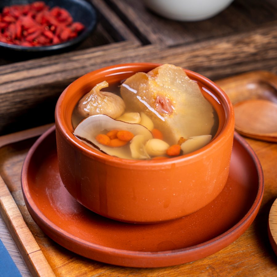 A red bowl of pear, dried fig, lily bulb, lotus seed and wolfberry soup on a wooden tray, with other traditional Chinese ingredients around it.