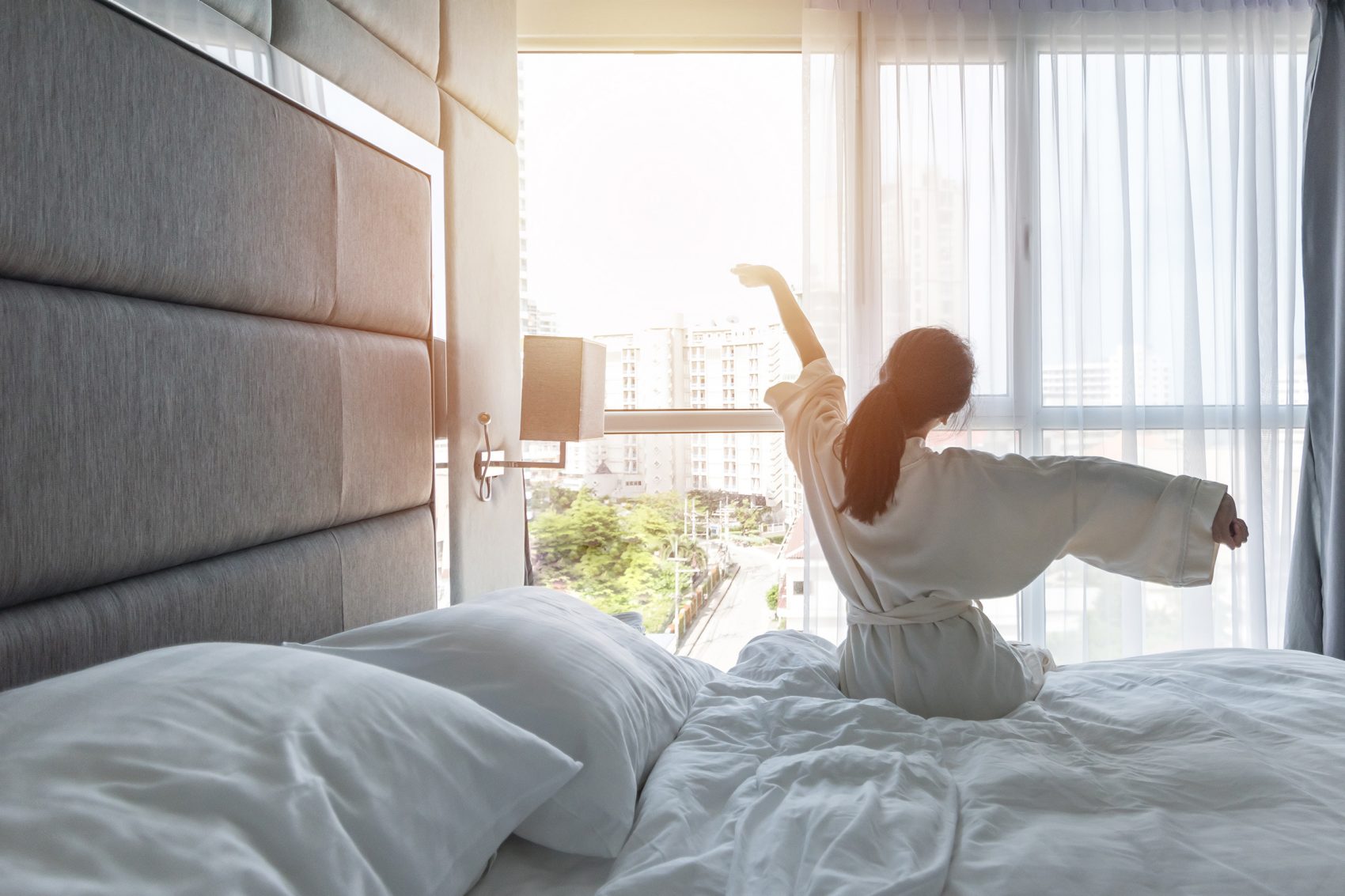 A woman stretches her arms as she gets out of bed, the sunlight streaming in through the windows. 