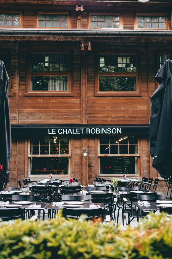Outdoor dining tables are arranged on a patio in front of a wooden cabin-style building. A sign on the building reads “Le Chalet Robinson”. 