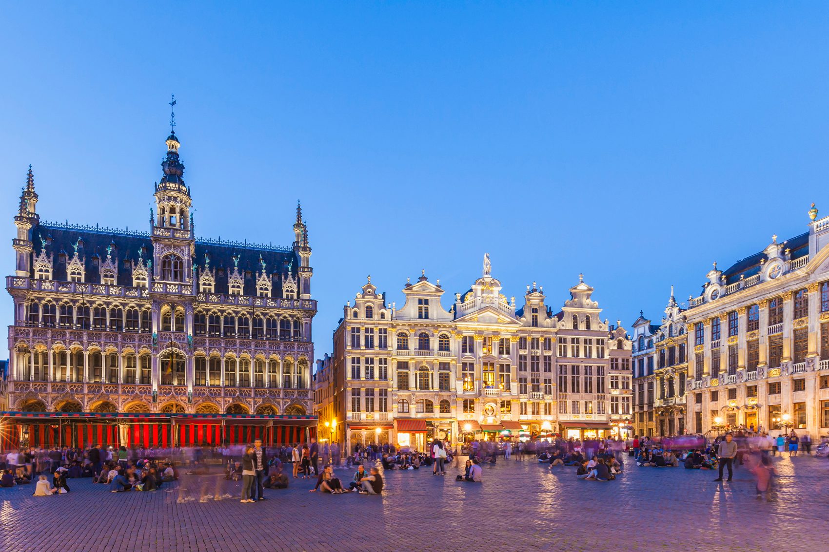 The large square of Grand Place in the evening. The ornate buildings that line the square are lit by warm lights. People are sitting or walking around the square.  