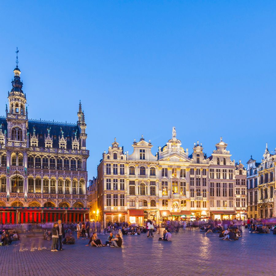 The large square of Grand Place in the evening. The ornate buildings that line the square are lit by warm lights. People are sitting or walking around the square.  
