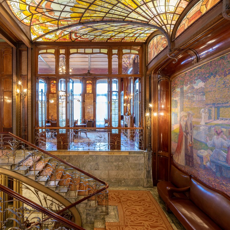 The Art Nouveau lobby of Hotel Solvay. The room features a stained-glass ceiling, a staircase with wrought iron bannisters and a mosaic mural.