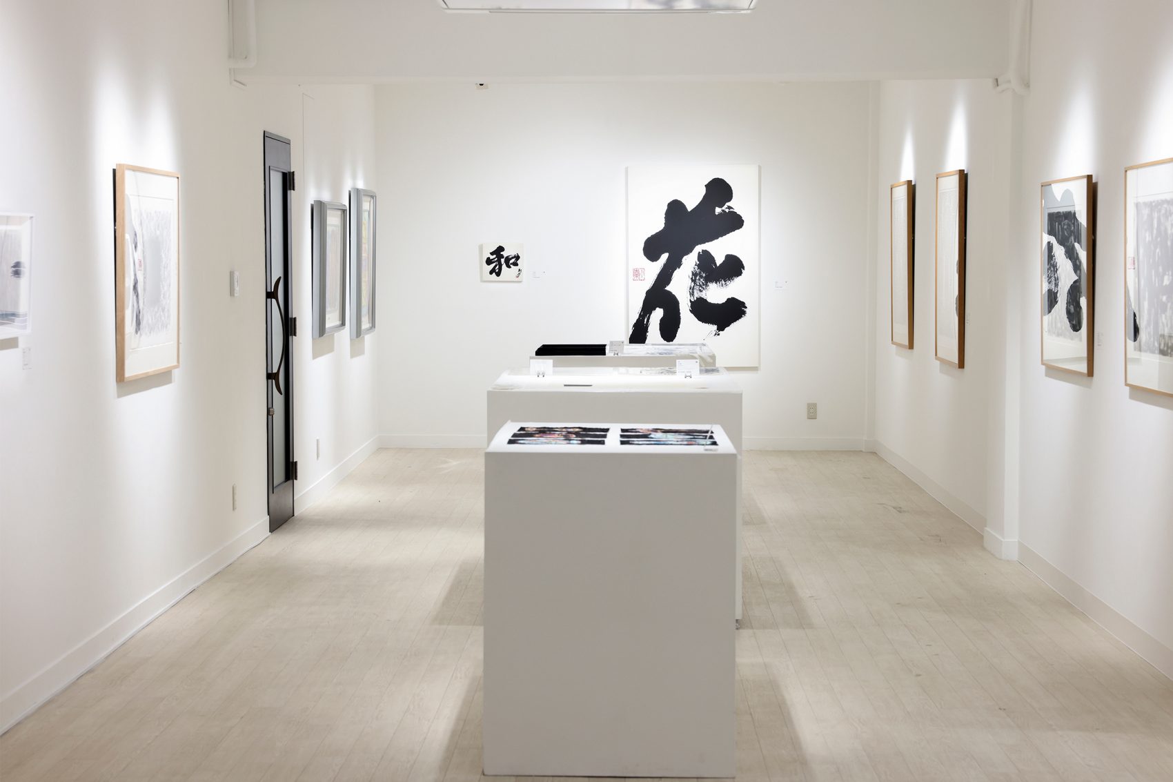 An all-white gallery room with two pedestals in the centre of the room; framed paintings are displayed on the walls and on the pedestals.