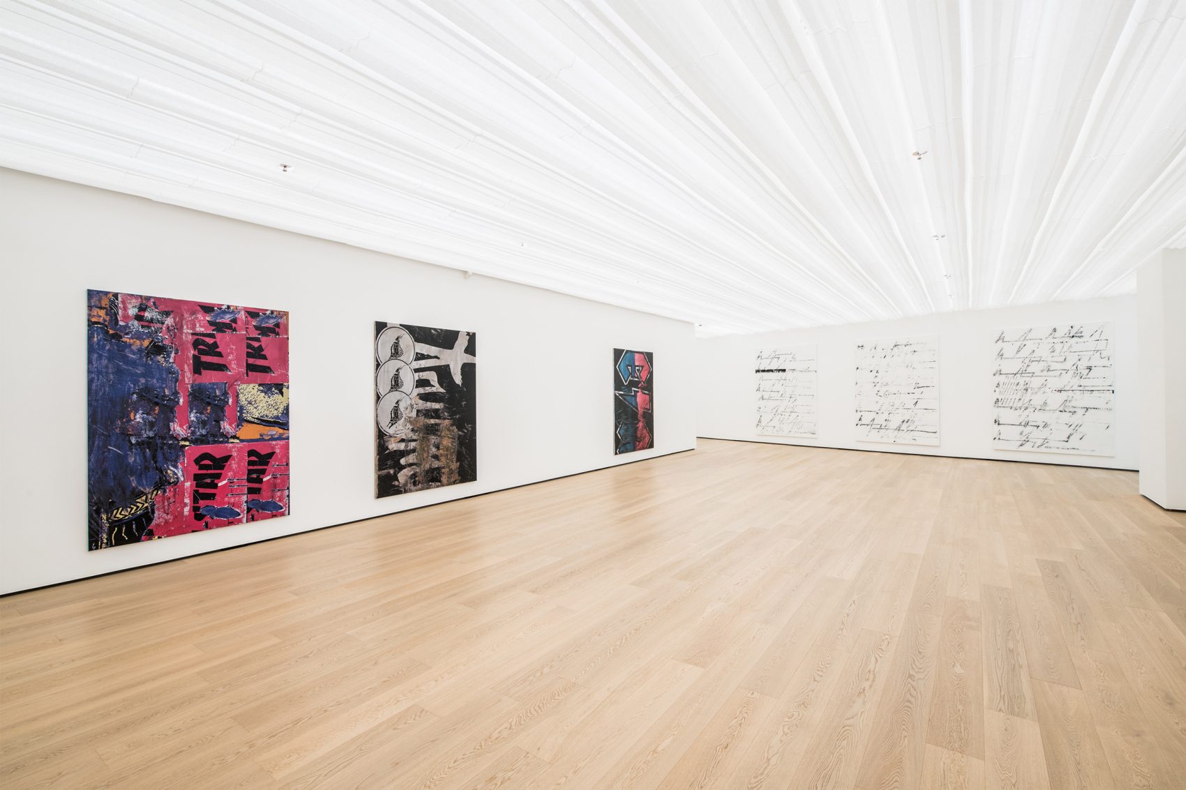 Wide view of an empty gallery; it is brightly lit, with several large canvases displayed on the white walls.