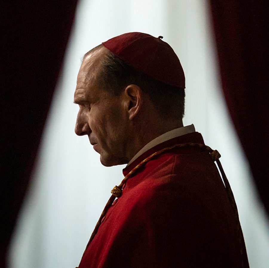 In a scene from the film Conclave, lead character Lawrence (Ralph Fiennes) is dressed in red cardinal robes, in a dark room illuminated by a streak of light.