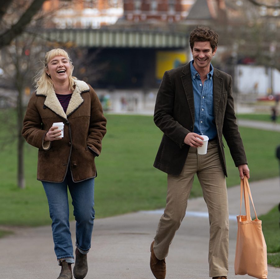 In a scene from the film We Live in Time, couple Almut (Florence Pugh) and Tobias (Andrew Garfield) walk through a London park on a winter day, holding cups of coffee and laughing. 