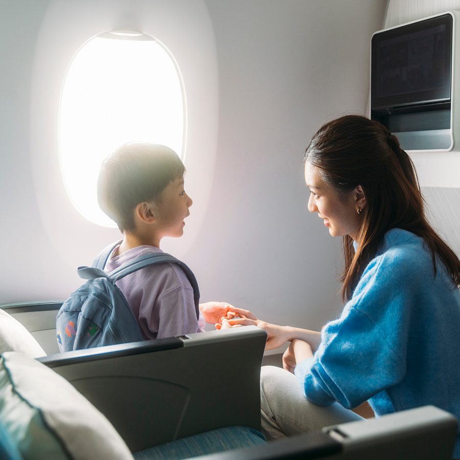 A woman and young boy smiling and holding hands sitting by the window inside a Cathay Pacific aircraft.