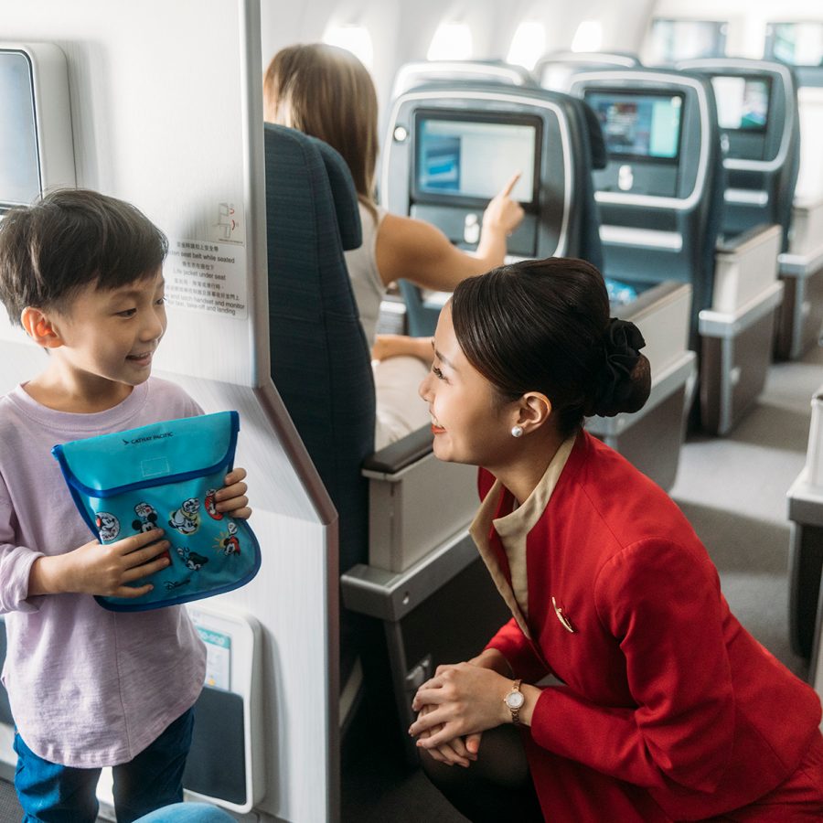 A Cathay Pacific air hostess happily talking to a young boy as he holds a bag of Cathay Pacific toy set inside an aircraft. 