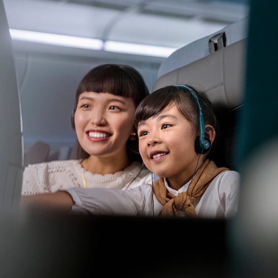 A young girl and her mother wearing a pair of Cathay Pacific headset while happily watching in-flight entertainment.