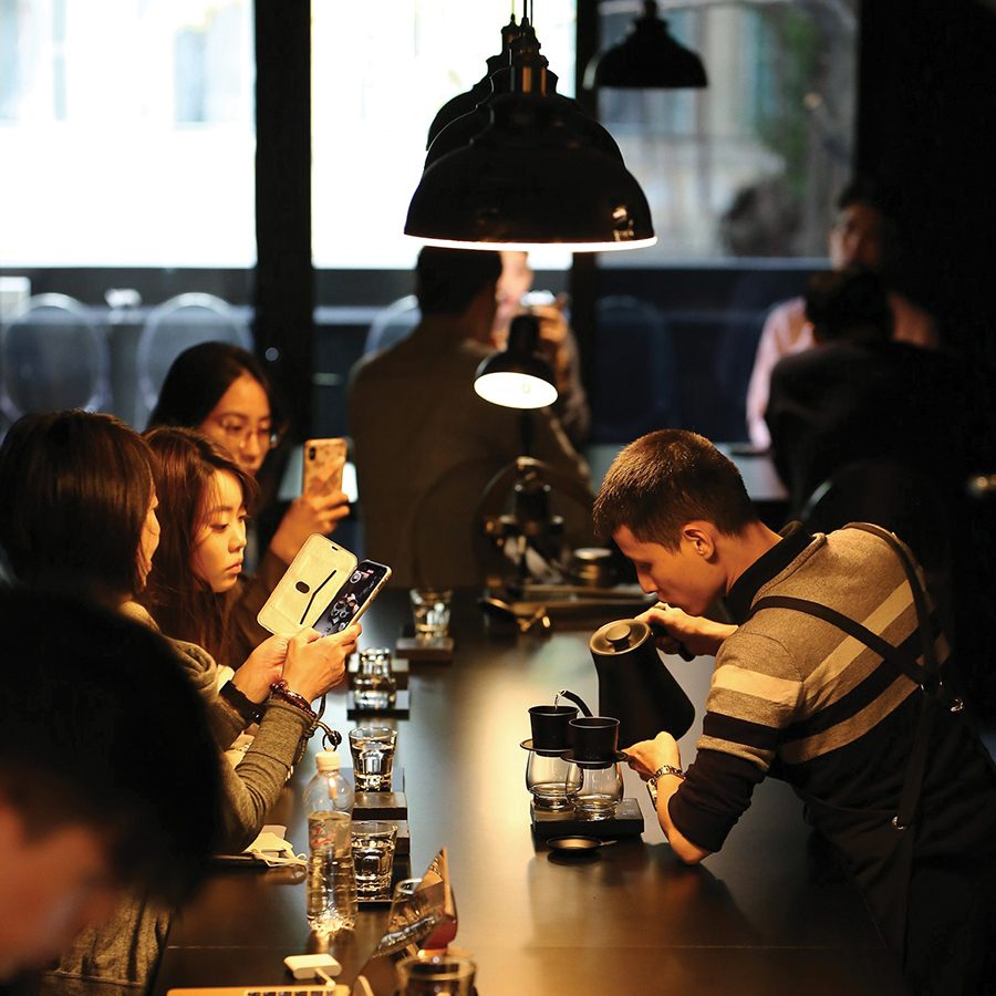 Customers sit at a counter and take videos of a barista making hand-poured filter coffee at Phin Bar by Refined, Hanoi. 