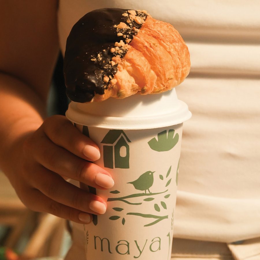 A closeup of a woman holding a Maya Bakery & Bar paper cup with a vegan chocolate croissant sitting on top.