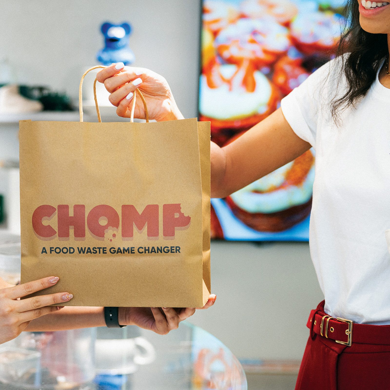 A smiling woman picks up a paper bag containing a Chomp mystery meal from Cookie DPT.