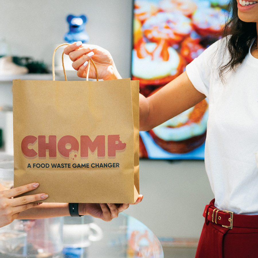 A smiling woman picks up a paper bag containing a Chomp mystery meal from Cookie DPT.