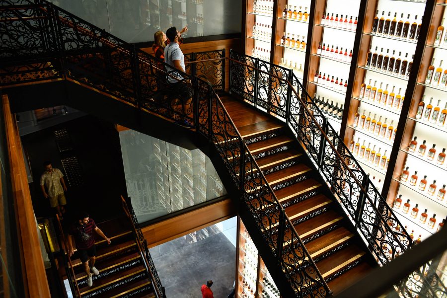 A woman and a man stand on the staircase inside the Sazerac House looking at a wall with shelves of liquor bottles.