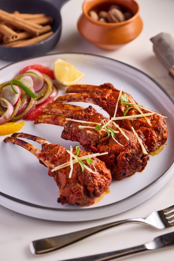 A closeup of a plate of tandoori lamb chops with lemon and vegetables.