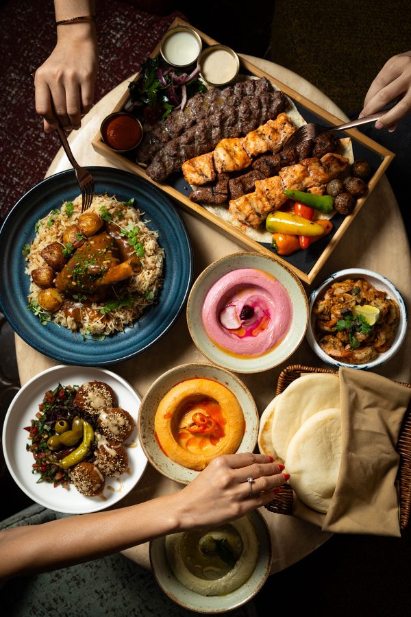 An overhead family style shot of people reaching for food, featuring various middle eastern dishes, pita breads, hummus and halal meats served at halal restaurant Bushra in Tsim Sha Tsui.
