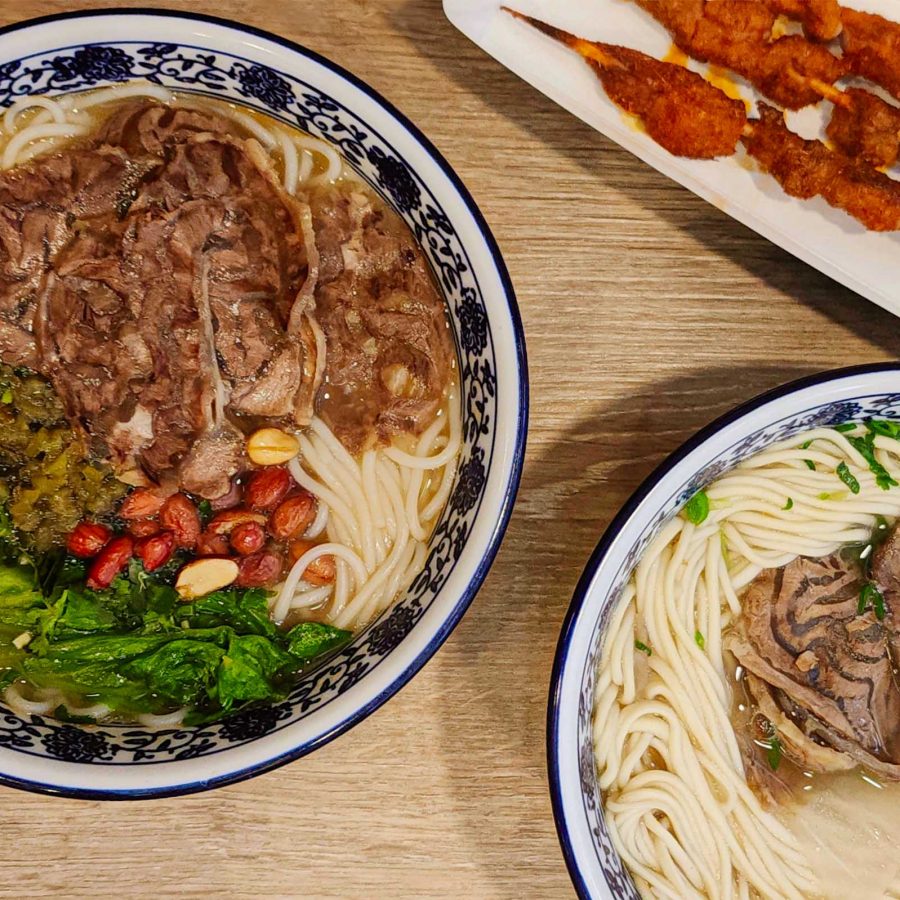 An overhead shot of two bowls of hand-pulled noodles with halal braised beef slices and cumin-spiced lamb skewers.