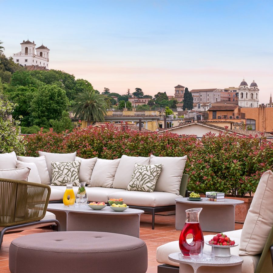 An outdoor view of the Hotel de Russie rooftop decorated with plush outdoor lounge sofas, green trees and flowering shrubs, with a city view of Rome in the background.