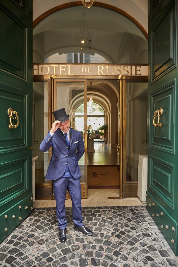 A hotel doorman in blue suit and top hat cheerfully greeting outside the green doors of Hotel de Russie in Rome.