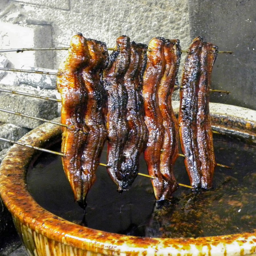 Four pieces of grilled eel, held up on skewers, are dipped into a vat of sweet sauce at Atsuta Horaiken.