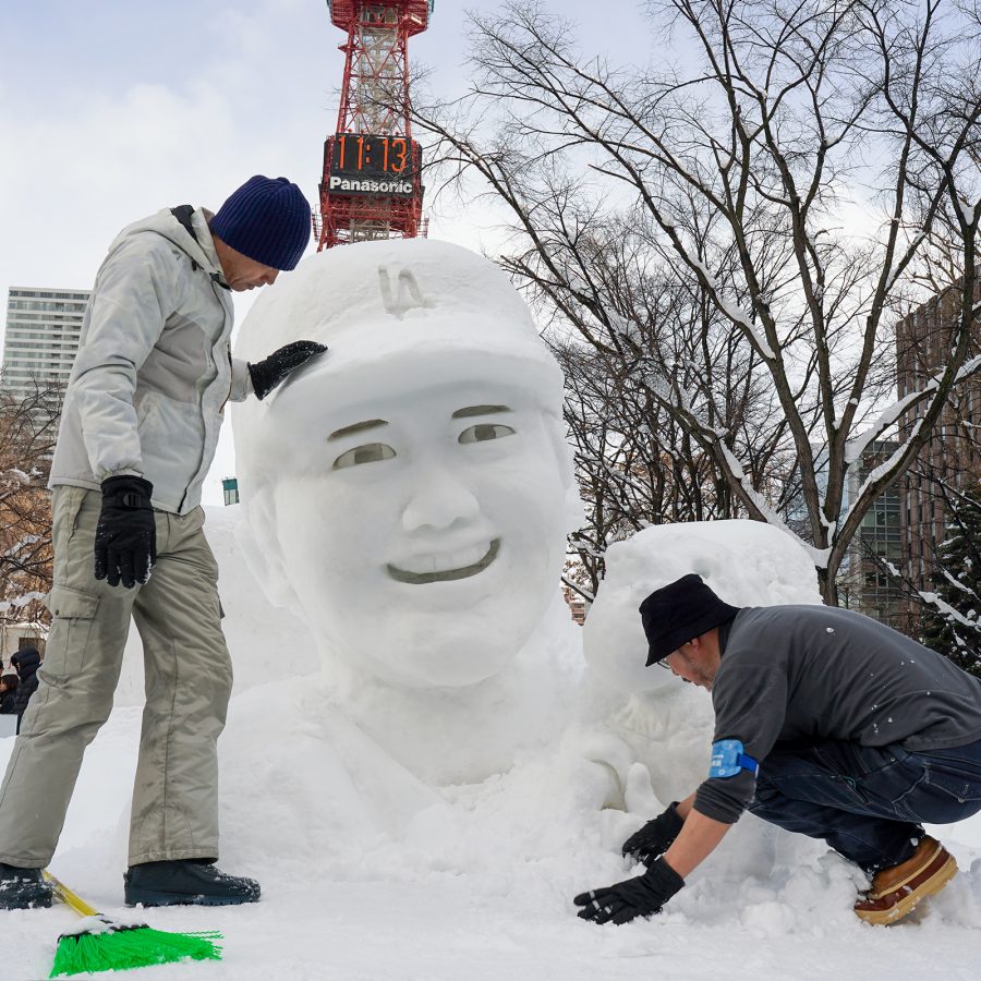 Two people work on a snow sculpture outdoors in Odori Park as the Sapporo TV Tower pokes into the sky in the background.   