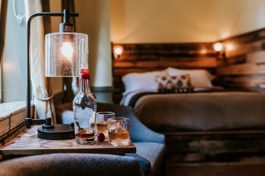 A bottle of bourbon poured into two small glasses with a view of the bed in the background at The Samuels House.