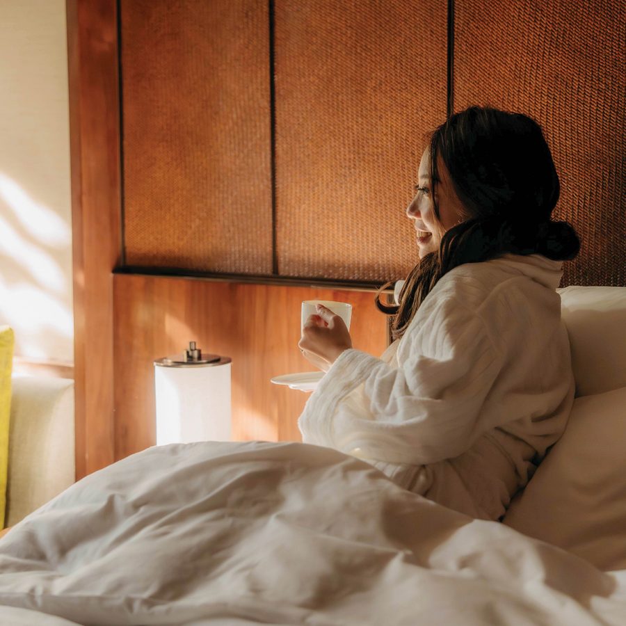 An Asian woman sitting in a hotel bed, holding a cup of coffee and smiling toward the window