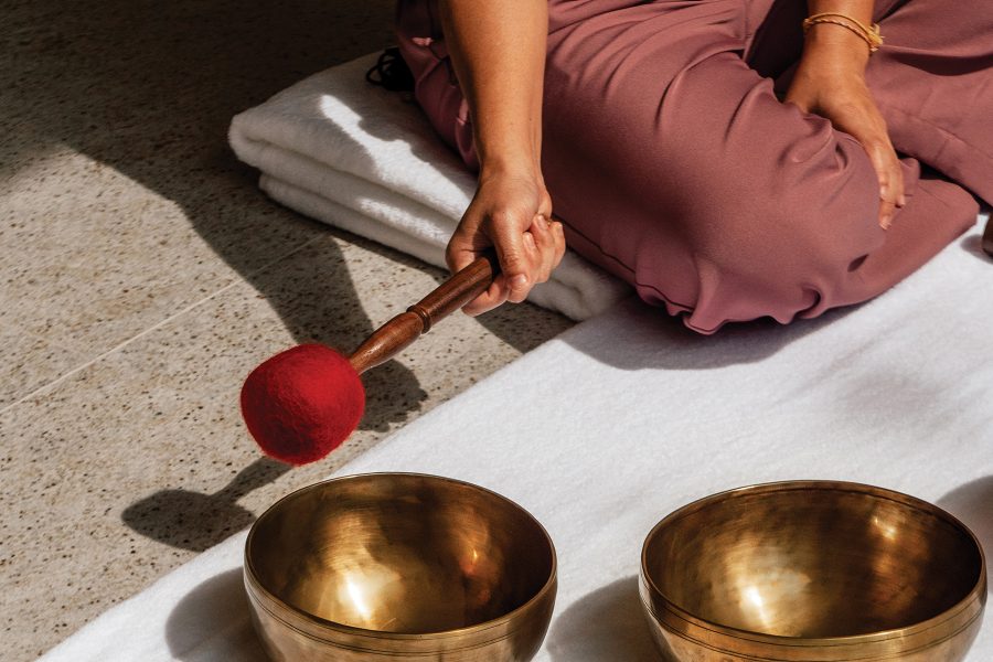 A woman sits on a towel on the floor, holding a mallet ready to strike a pair of singing bowls, with sunlight streaming in
