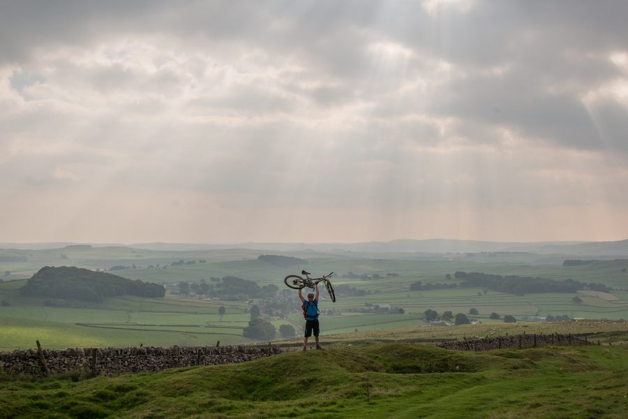A man holds a bike in the air in the English countryside.