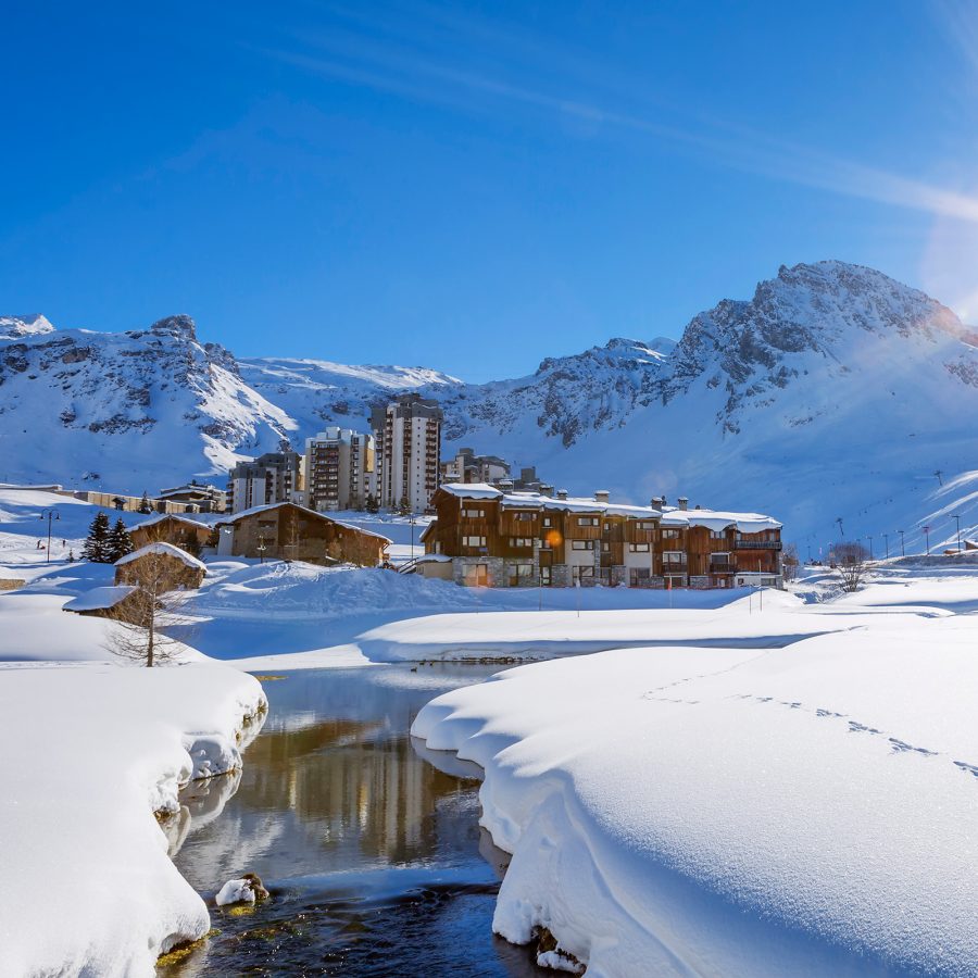Snow topped cabins in a French alpines resort shine in the sunlight under a blue sky.