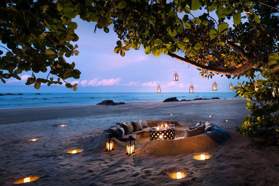 An intimate set-up on a beach with a large tree over a fire pit and pendant lamps; an example of the Designer Dining by Anantara service offered by Anantara Desaru Coast Resort & Villas. 