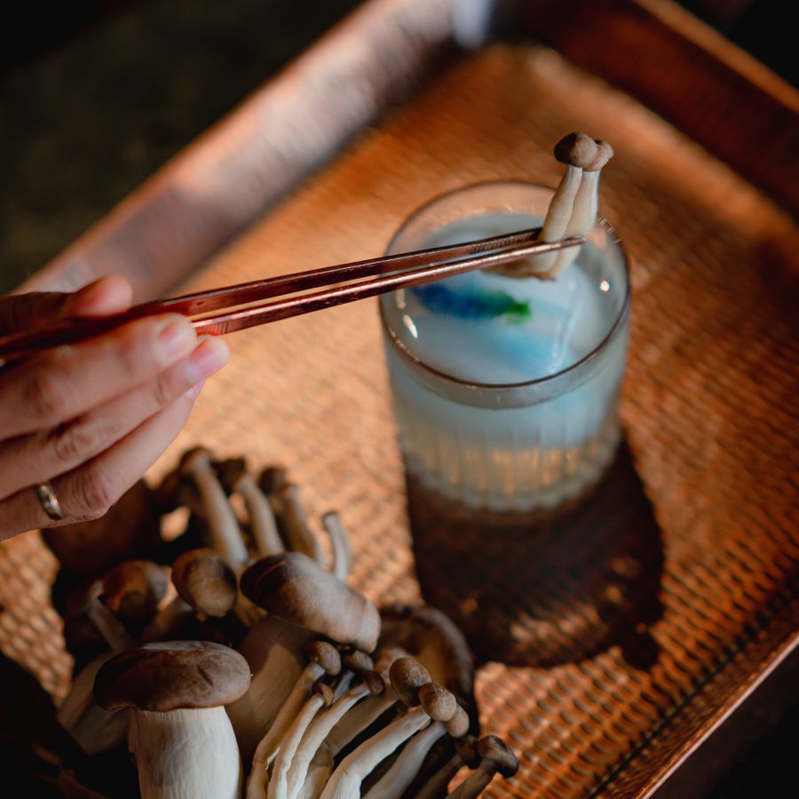 A hand holding mushrooms with forceps over a cocktail at Bambu Indah, Bali. 