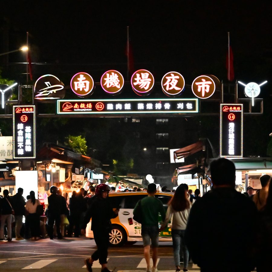 A labelled archway marks the location of Nanjichang Night Market as people and cars pass by it on the street. 