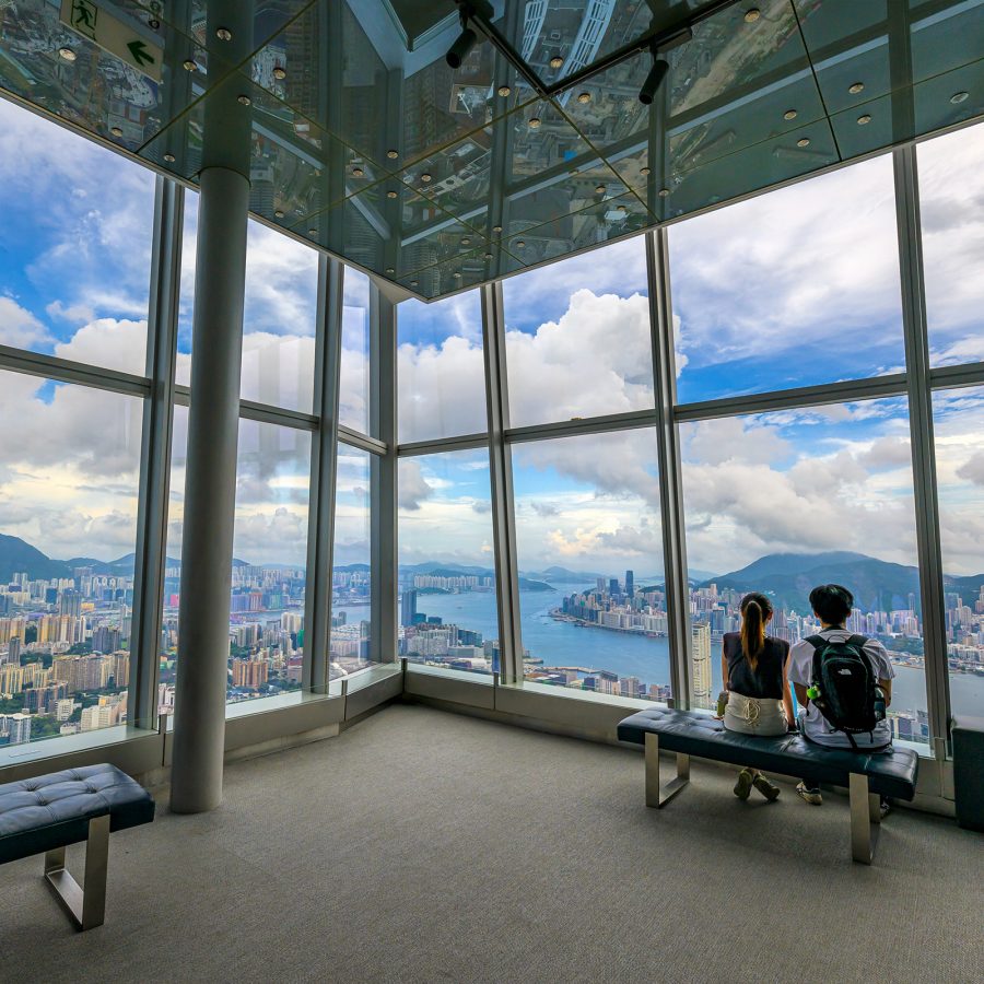 A couple of man and woman sitting on a bench looking out to panoramic view of Hong Kong’s skyline from Sky100 in the top of International Commerce Centre.