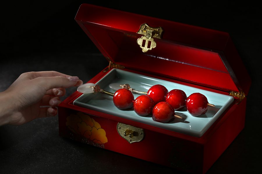 A closeup of a hand picking up a candied dessert from a luxurious red jewellery box, served at Chinesology contemporary Chinese restaurant in IFC, Hong Kong. 