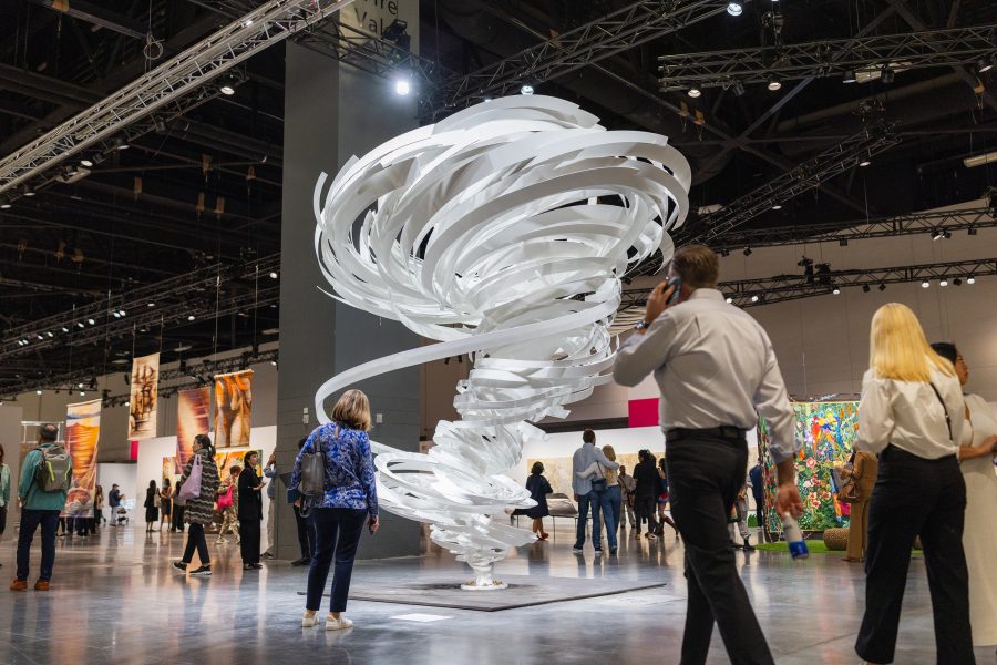 People walking on the show floor of Art Basel Miami Beach 2024. Alice Aycock’s Goya, a large-scale sculpture resembling twisting white ribbons, is in the middle of the shot. 