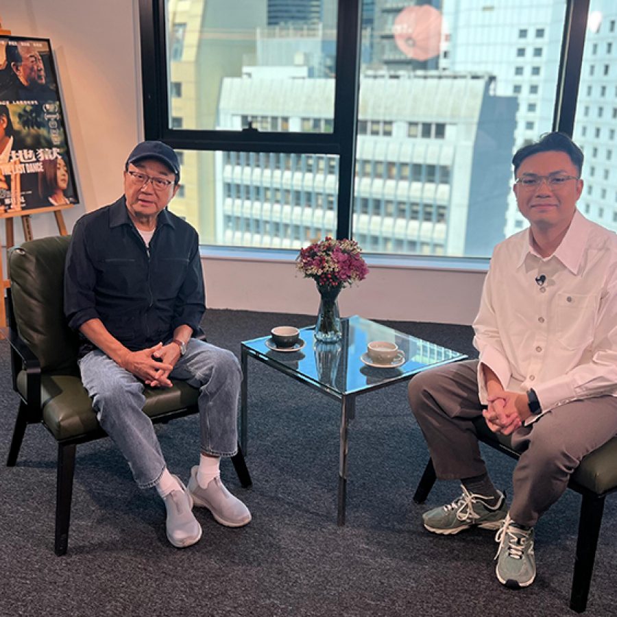 Actor Michael Hui sits on a low chair in a bright room; opposite him sits an interviewer. Behind his is a poster for the film The Last Dance. 