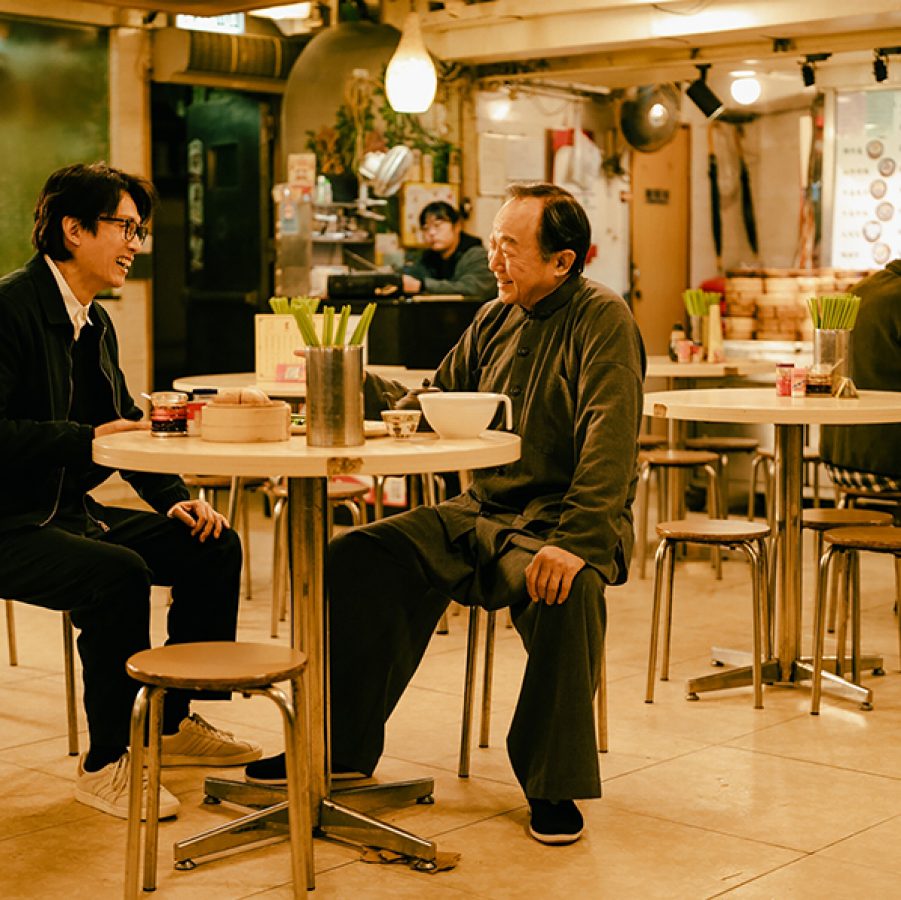 Dominic (Dayo Wong) and elderly priest Master Man (Michael Hui) laugh while sitting on stools at a table in a local restaurant, in a scene from The Last Dance.