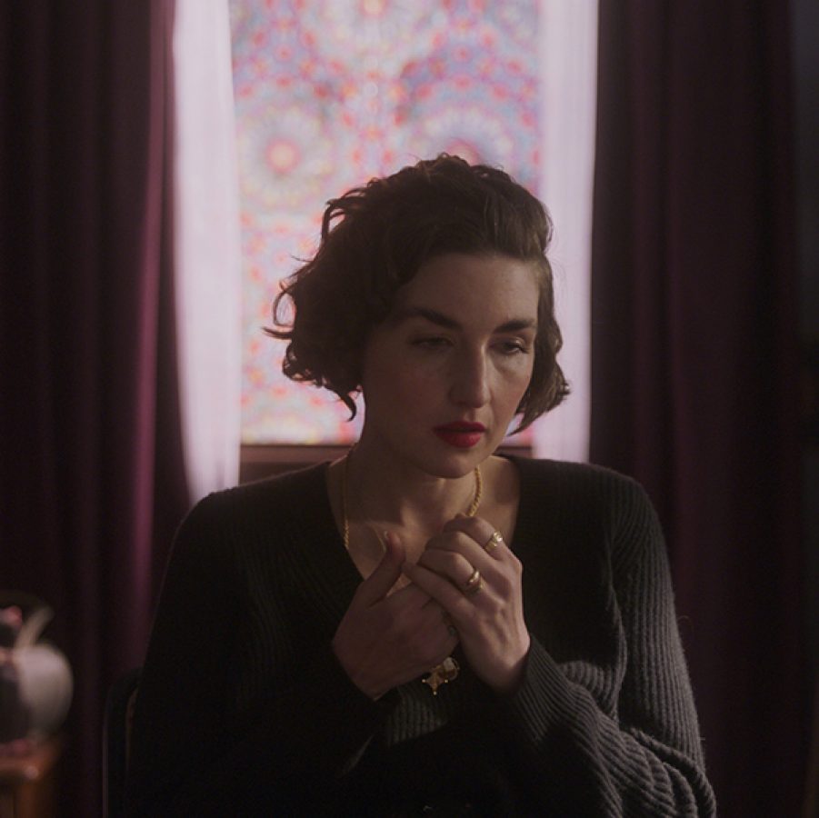 Filmmaker Elizabeth Sankey sits on a chair in a softly lit bedroom in a scene from Witches. 