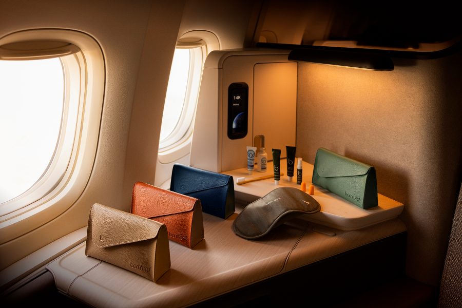 A lifestyle shot of the new Cathay Pacific Bamford amenity kits in four colours next to an aircraft window in the Aria suite of the Business cabin